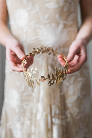 Model in white gown holding a gold laurel leaf bridal headpice